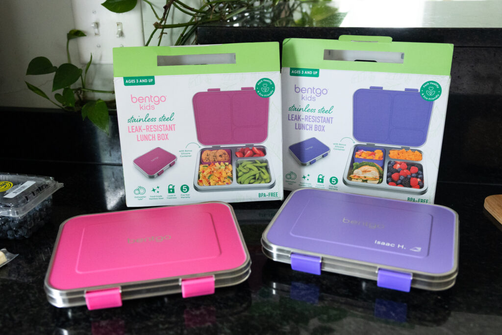 Two Bentgo Kids stainless steel leak-resistant lunch boxes in pink and purple on a kitchen counter, shown with their packaging boxes featuring BPA-free and eco-friendly labels, photographed in Raleigh NC documentary family style