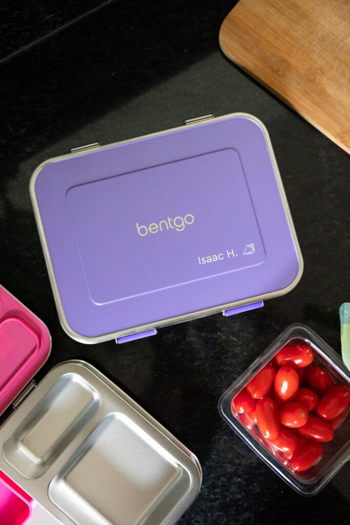 Bentgo kids stainless steel lunch box in purple with name personalization including Isaac H. and a seashell design, next to open pink stainless steel lunch box and an open plastic carton of grape tomatoes