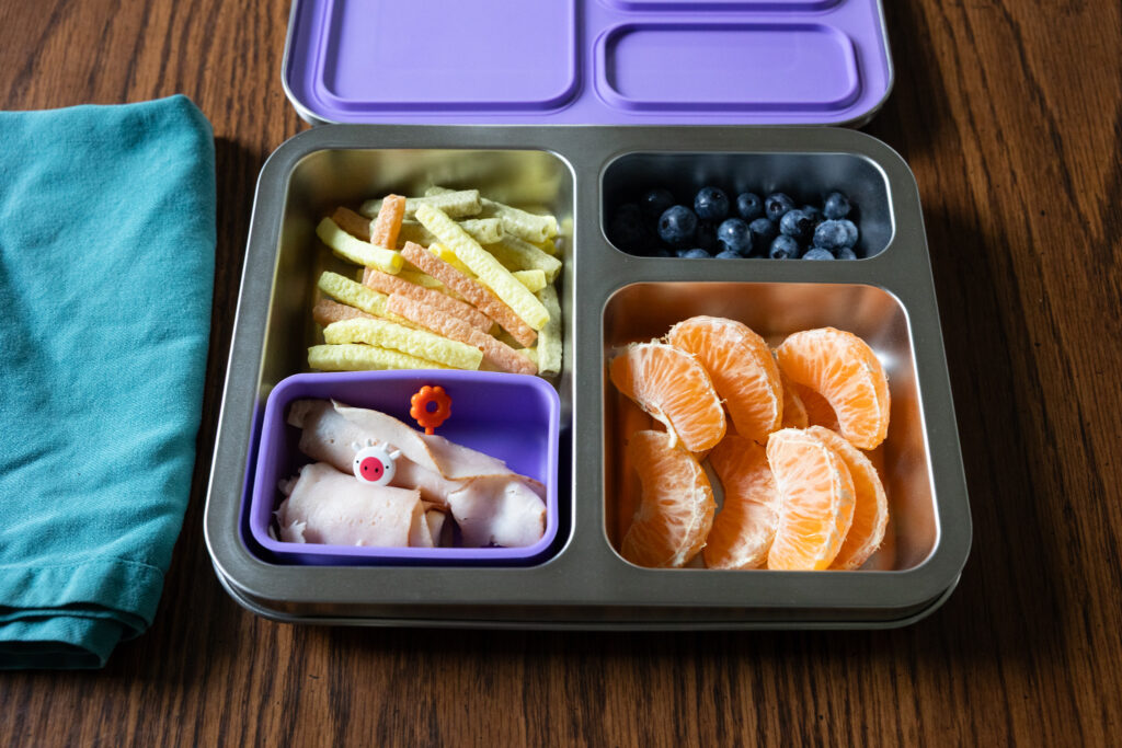 Open purple-lidded Bentgo kids stainless steel lunch box on a wooden table with a teal cloth napkin to its left. The lunch box contains veggie straws, lunch meat & cheese roll-ups with kid-friendly plastic toothpicks, blueberries, and orange slices, photographed by Light Creative in Raleigh NC
