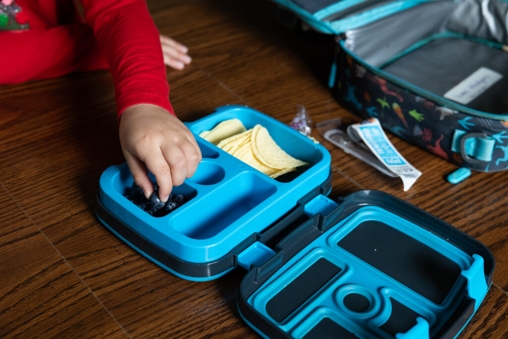 Child in red sleeve picking blueberries from blue Bentgo Kids lunch box with chips and snacks on wooden table, candid Raleigh NC documentary family photography