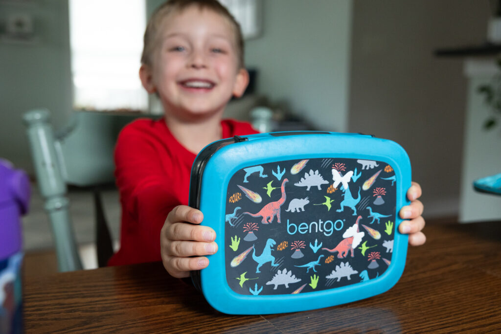 Six-year-old boy smiles while holding the leak-proof plastic Bentgo Kids Prints Lunch box that he has used for the past four years through preschool. The lunch box has a blue border with a dinosaur design on the top.