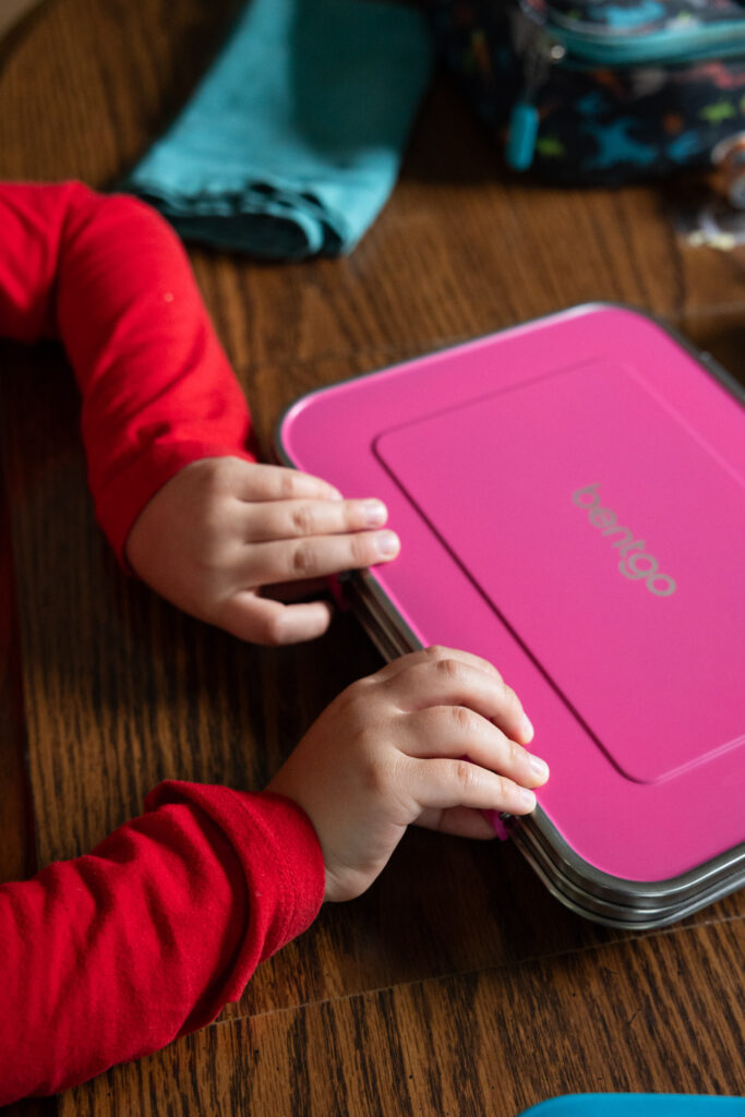 Photo 1 in a 3-photo series of a child wearing a red shirt opening a pink stainless steel Bentgo kids lunch box. Close-up of his hands as he easily opens the kid-friendly latches, photographed by Raleigh family photographer.
