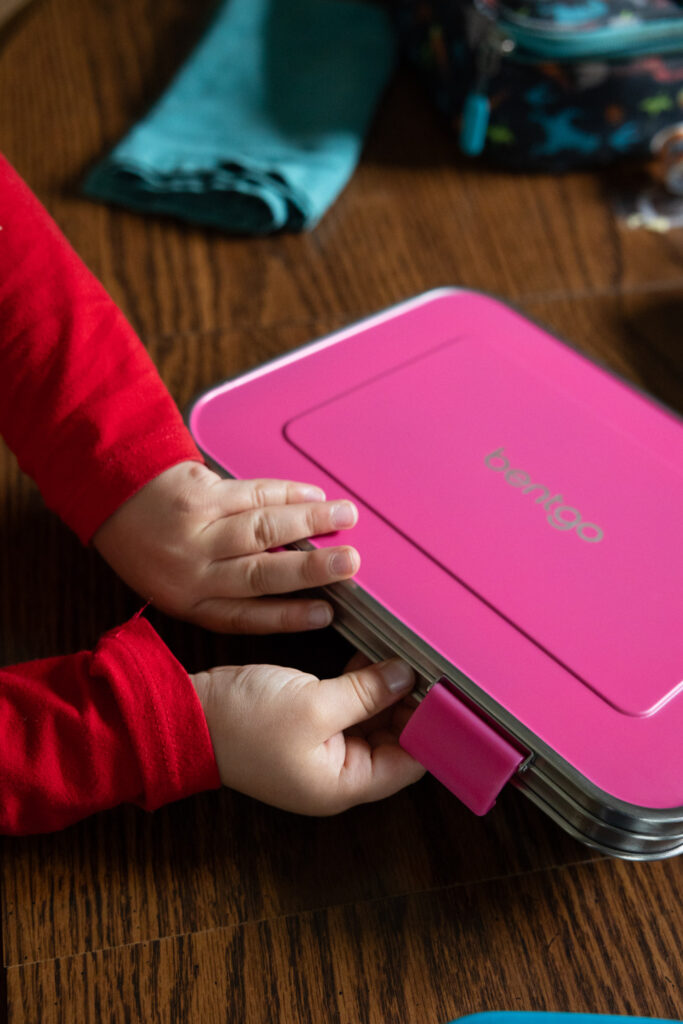 Photo 2 in a 3-photo series of a child wearing a red shirt opening a pink stainless steel Bentgo kids lunch box. Close-up of his hands as he easily opens the kid-friendly latches, photographed by Raleigh candid family product photographer.