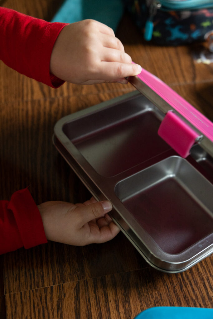 Photo 3 in a 3-photo series of a child wearing a red shirt opening a pink stainless steel Bentgo kids lunch box. Close-up of his hands as he easily opens the kid-friendly latches, now with the lunch box fully open, photographed by Raleigh candid family photographer Light Creative.