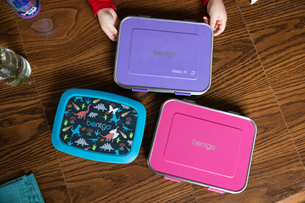 Overhead view of three colorful Bentgo Kids lunch boxes in purple, pink, and blue-bordered dinosaur print on wooden table, Raleigh NC documentary family product photography