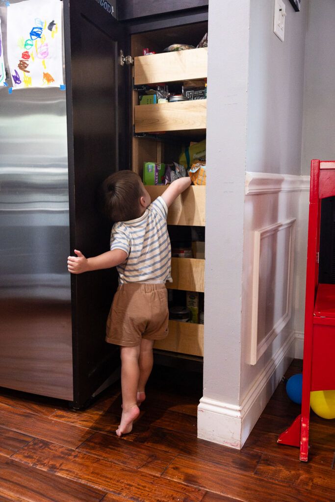 In-home editorial family photography - a 3-year-old boy stands on tip-toes and holds onto the pantry cabinet door as he reaches in to find the snack he wants
