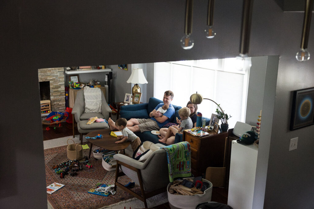 Candid documentary family and newborn photography in Raleigh capturing real life as it is right now. The camera looks in on a family's living room while toys are strewn about, with parents, newborn, and eldest brother sit on the couch and the middle brother leans over the coffee table.