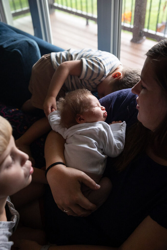 in-home candid documentary newborn photography with siblings and parents - mom holds newborn baby girl while baby's middle brother holds her hand and her oldest brother leans in