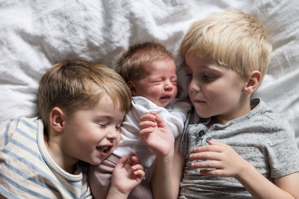 Raleigh natural newborn photography with brothers - two brothers laying on a white blanket and snuggling with newborn baby sister in between