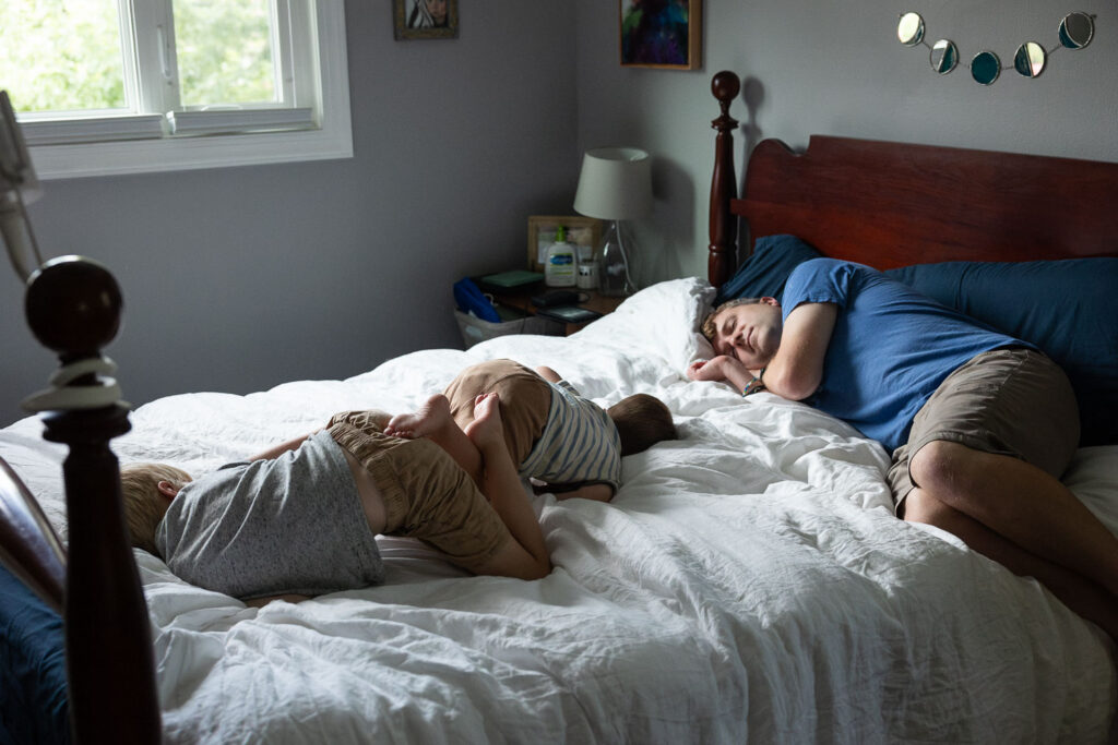 Photo inspiring "Poem of the Tangled Feet (965) shows a dad laying on a queen-sized bed with two boys with their backs facing up, bare feet and legs tangled up in each other after rough-housing.