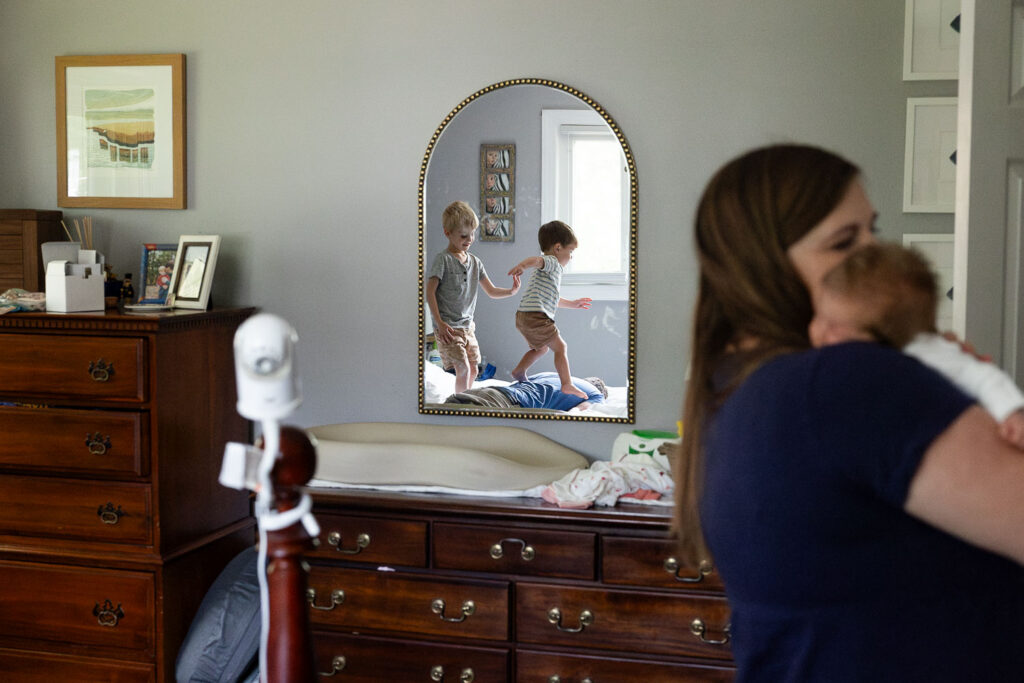 Natural in-home newborn photo showing mom holding baby in the foreground and a mirror reflection of the baby's brothers jumping on their dad's back while he lays face-down on a bed
