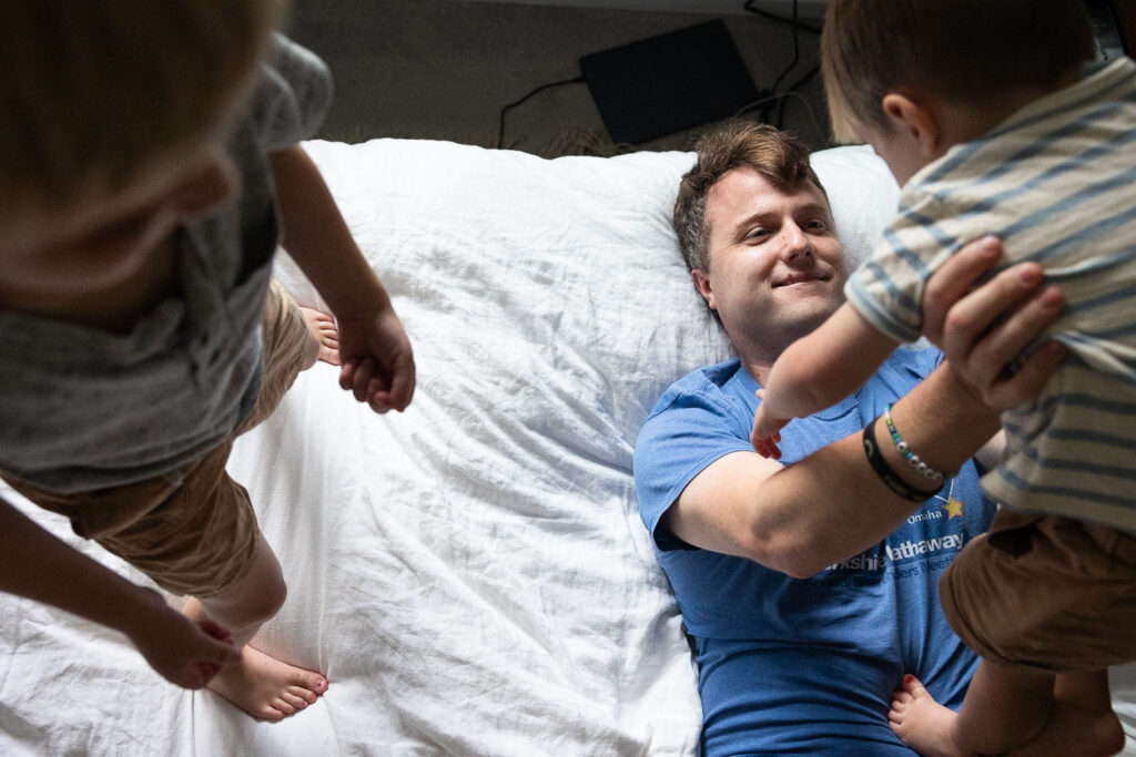 Candid family photo with dad in a t-shirt laying on his back on his bed, holding his toddler son up above him while the older brother stands beside and watches