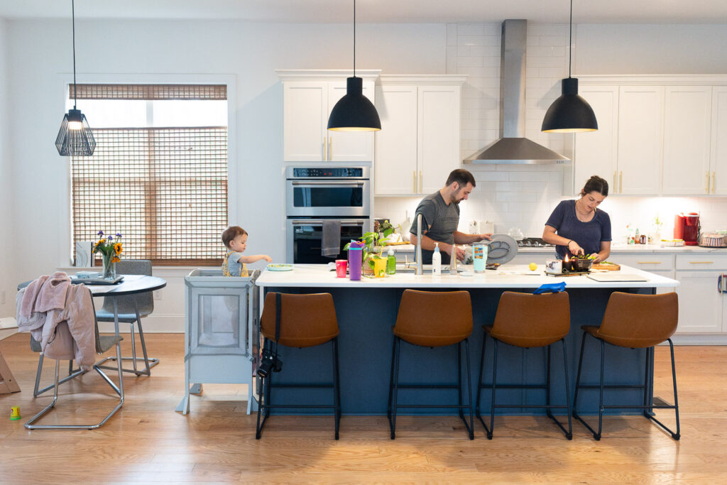 South Raleigh family makes dinner in the kitchen, with dad washing dishes, mom cutting food, and toddler standing in helper stool on the other end of the counter and eating from a plate