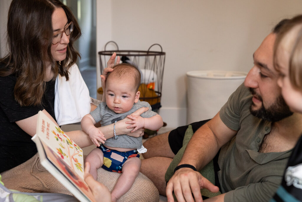 Downtown Raleigh candid documentary family photo session, mom smooths her finger over her baby's soft, downy hair while dad lays on his side and reads a book to their toddler