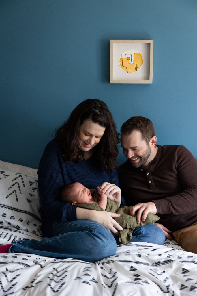 Happy parents holding and gazing at their newborn son while sitting on their bed during Raleigh in-home newborn session