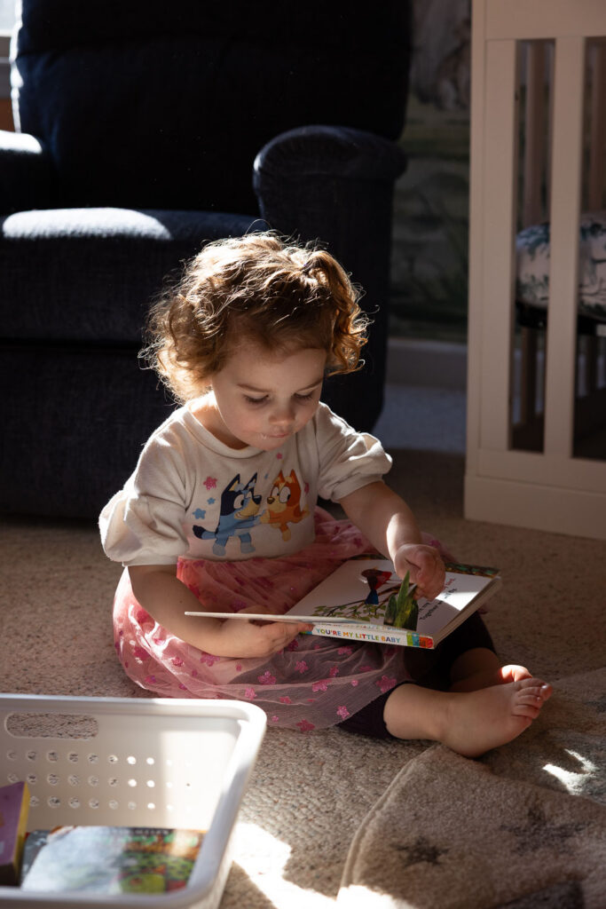 Raleigh candid documentary family photos, toddler girl reads a board book with direct light in the nursery