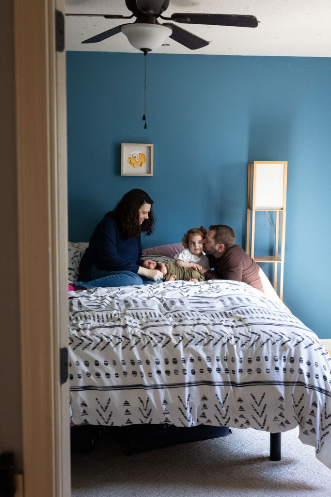 Raleigh in-home newborn photo session, looking into parents' bedroom with baby lying on mom's lap and dad kissing toddler daughter's cheek