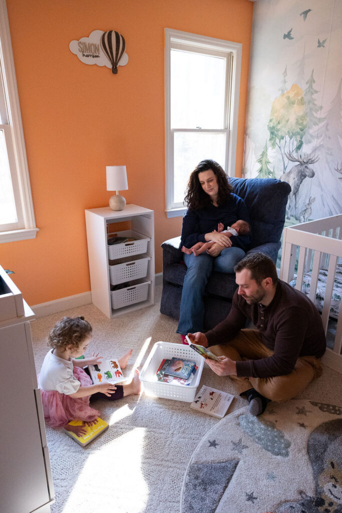 Newborn photos in Raleigh that show real life as family spends time in the nursery. Mom nurses the baby while dad and daughter read books while sitting on the floor.