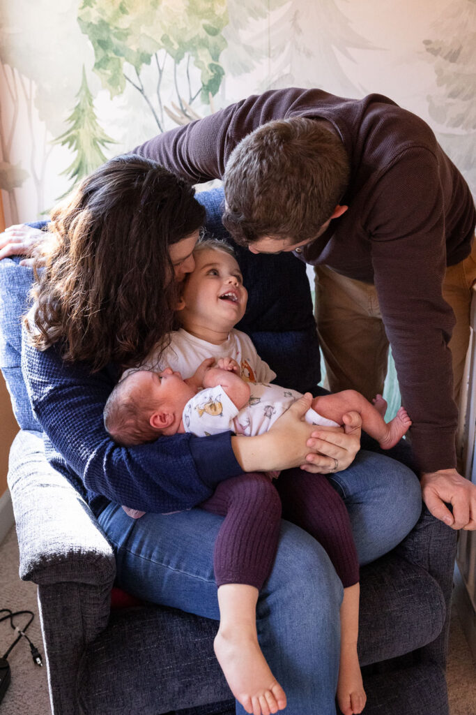 Mom sits in nursery glider with toddler daughter and newborn son while dad leans over the side of the glider to them during Raleigh newborn photo session