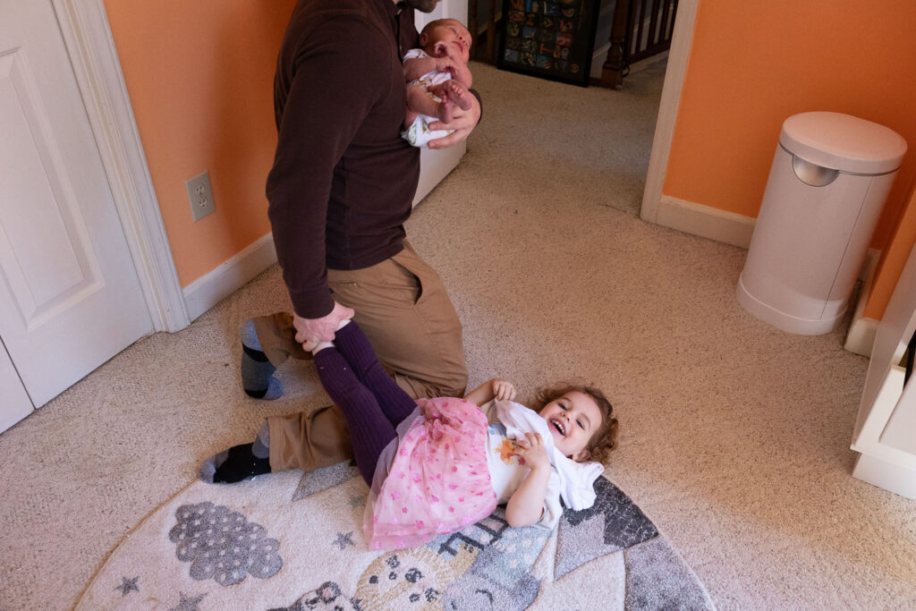 Candid family photo session in Raleigh, NC, dad stands on his knees and holds toddler's feet with one hand while holding newborn in other arm