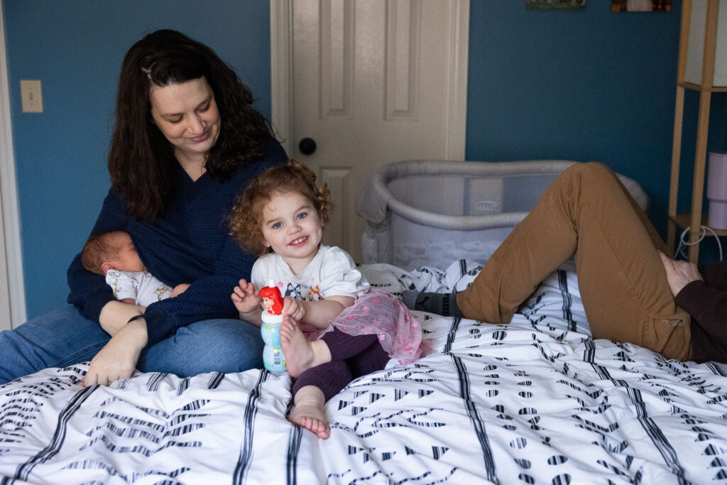 Family on parents' bed during Raleigh natural newborn photo session while mom nurses baby and toddler daughter smiles at camera