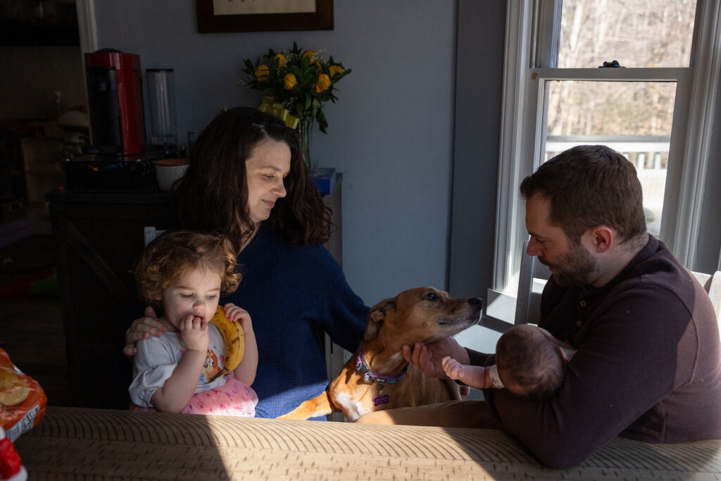 Candid newborn photo session that shows real life in Raleigh, family of four sits at kitchen table with their dog, toddler holding a banana like a phone, and dad holds newborn 