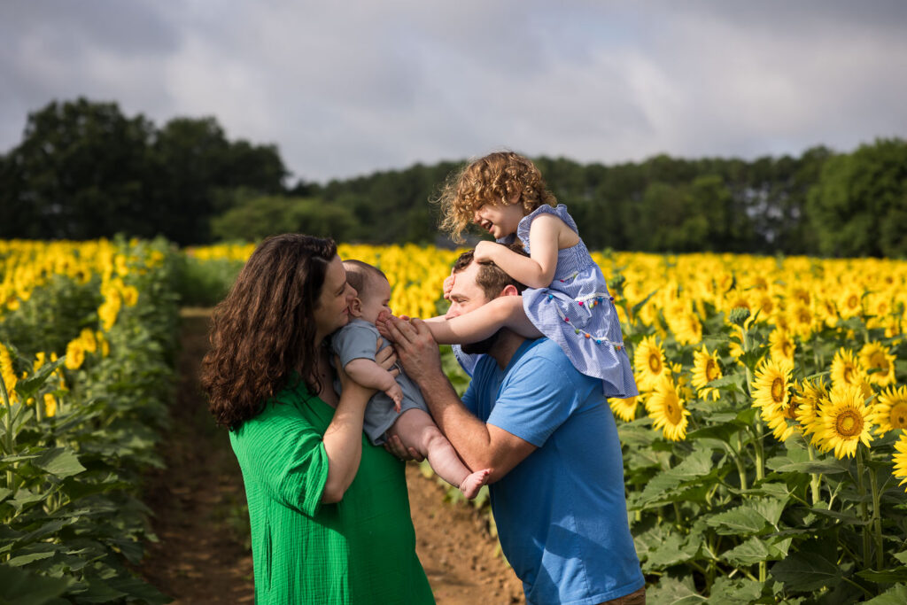 Playful family photos at Dorothea Dix Sunflower Field, daughter is on dad's shoulders and dad puts her feet on baby brother's cheeks, as he is held by mom