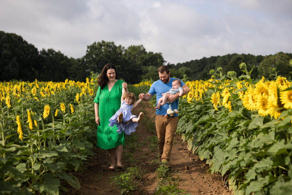Dix Park Sunflower family photo session, family walks toward camera while dad holds baby and mom and dad swing daughter between them