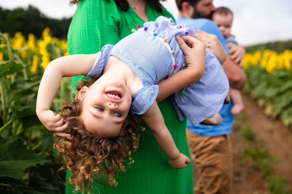 Dorothea Dix Park Sunflower Field photo sessions, toddler daughter is held by her mom and leans upside-down smiling with dad and baby in the background