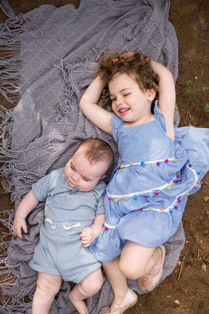 Toddler girl and baby brother sit and lay on a blanket in a row of sunflowers at Dorothea Dix Park in Raleigh