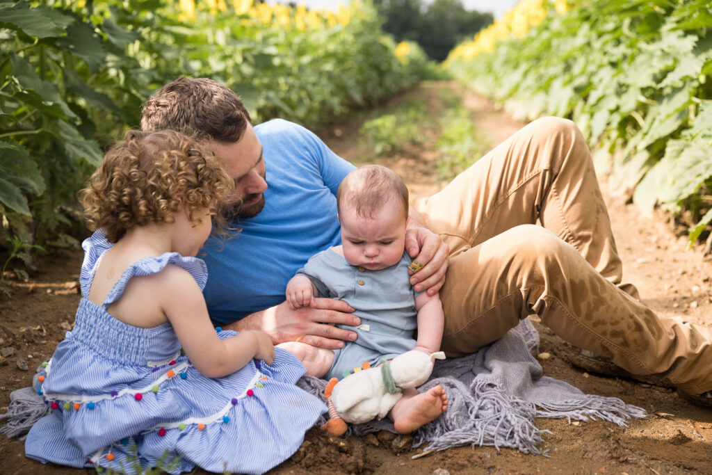 Dad, toddler daughter, and baby sit between rows of sunflowers at Dix Park photo session
