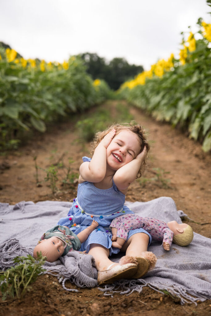 Toddler girl sits between rows of sunflowers at Dorothea Dix Park family photo session and smiles big while holding both sides of her head to keep her curly hair out of her face