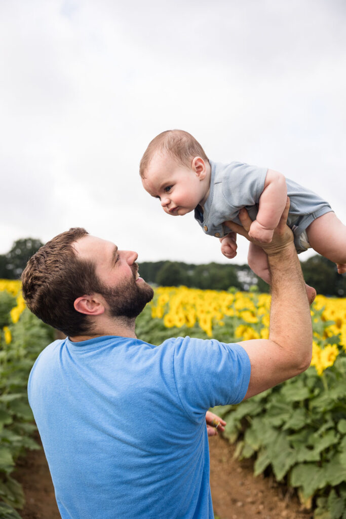 Dad holding 6-month-old baby up with one hand at Dix Park sunflower field photo session
