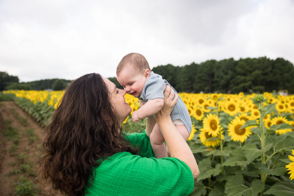 Mom holds 6-month-old baby son up and touches her nose to his with Dix Park sunflower field in the background