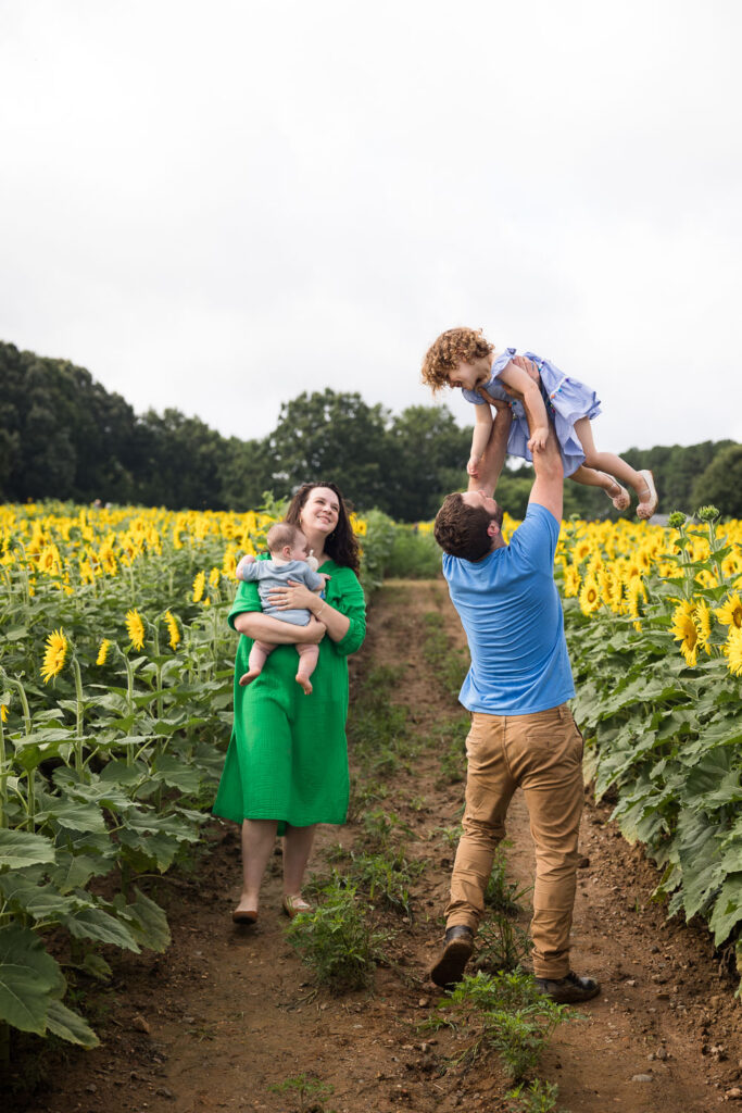 Baby milestone photo session at Dix Park sunflower field, mom holds baby and looks up at daughter as she is held up by the dad