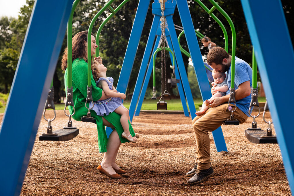 Family of four sits on tandem swings during baby milestone family photo session at Dix Park sunflower field