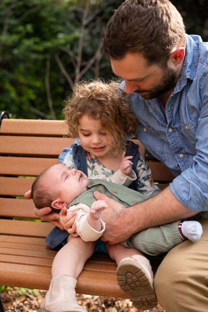 Raleigh baby milestone photo session, toddler girl holds baby brother on her lap with her dad on a park bench
