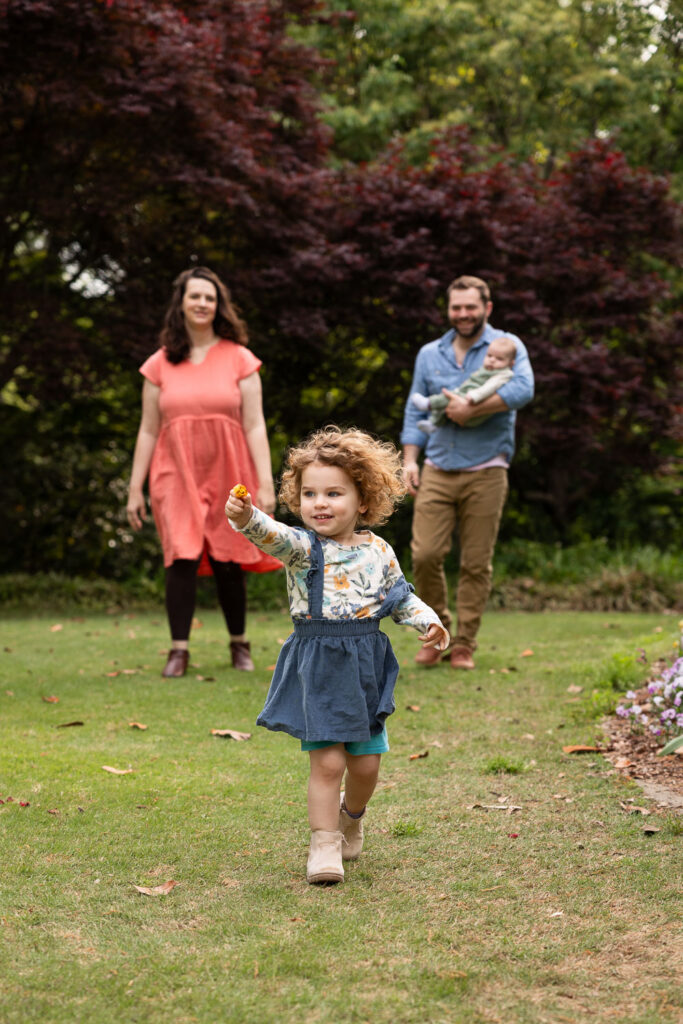 Fred Fletcher Park family photo session with toddler and baby, parents walk toward the camera with dad holding baby while toddler runs ahead holding a flower