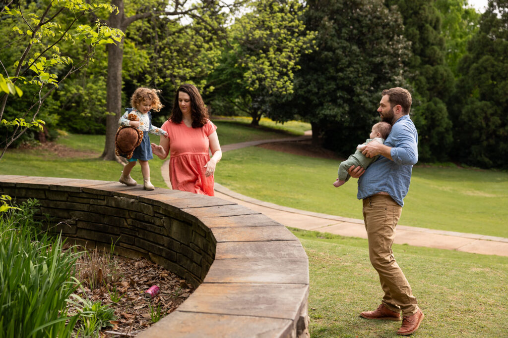 Fred Fletcher Park family photo session with toddler girl walking along short wall, mom supporting her, and dad looking on while holding baby
