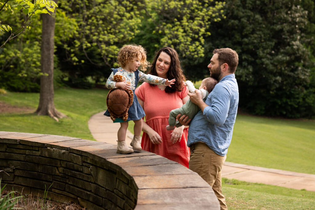 Fred Fletcher Park family photo session with toddler girl walking along short wall and reaching for baby brother as dad holds him, mom looks on