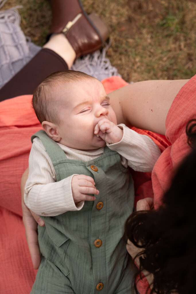 View from above of 3-month-old baby boy with his eyes closed as he is held by his mom and she sits on a blanket at his baby milestone photo session in Raleigh