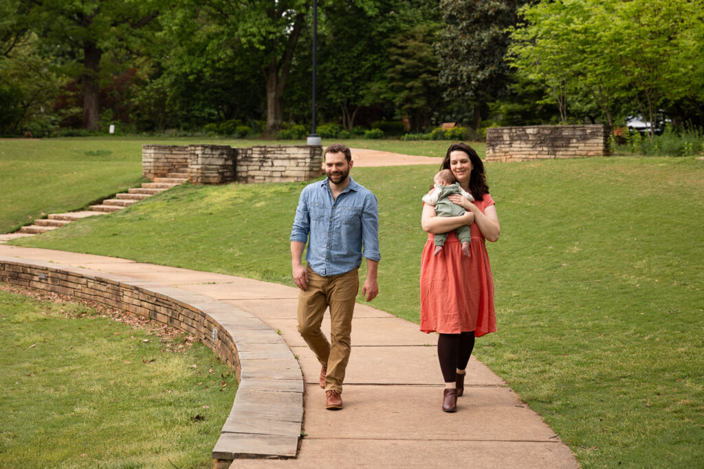 Baby milestone photo session in Raleigh at Fred Fletcher Park, parents walk along sidewalk toward camera while mom holds baby