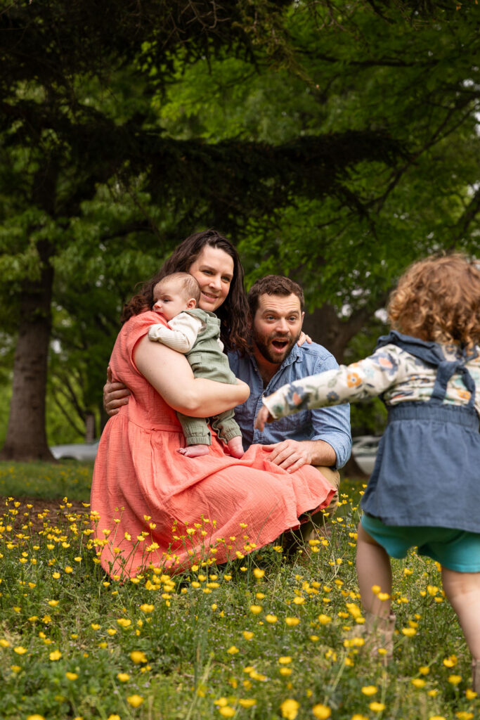 Family is in field of yellow flowers at Fred Fletcher Park in Raleigh, NC. Parents squat down while mom holds 3-month-old and toddler runs toward them away from the camera