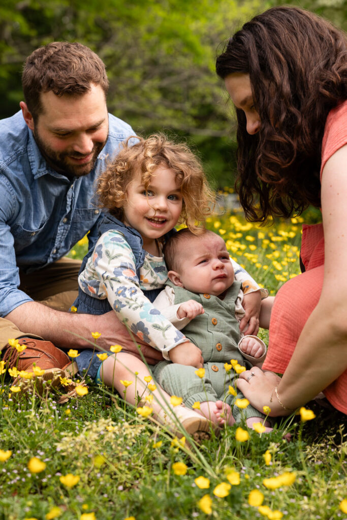 Toddler and baby brother sit in yellow flowers at Fred Fletcher Park while parents support them