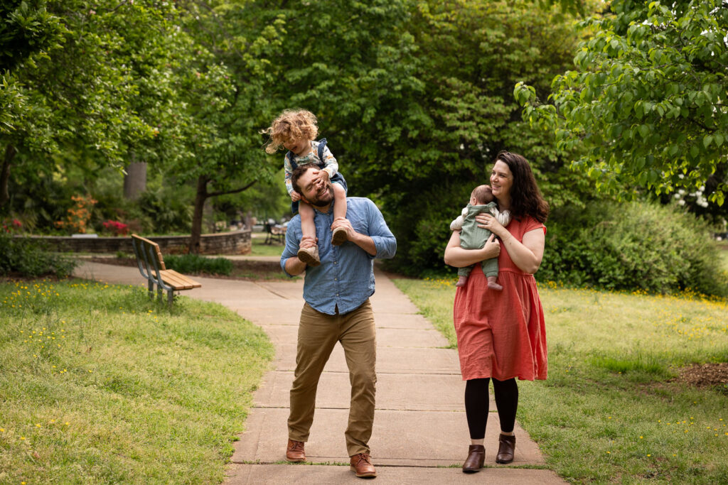 Fred Fletcher Park family photo session with baby and toddler, walking along sidewalk toward camera while mom holds baby and dad carries toddler on his shoulders