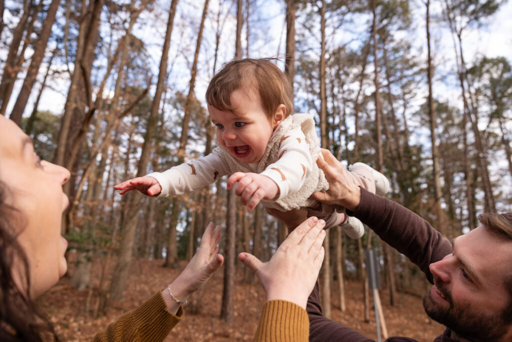 1-year-old baby milestone photo session at Lake Johnson Raleigh, dad helps smiling baby fly through the air to mom with outstretched hands
