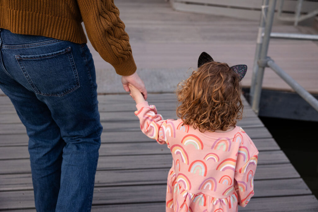 Toddler girl holds mom's hand while walking away from camera on Lake Johnson dock in Raleigh photo session