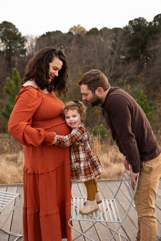 Raleigh maternity photo session at North Carolina Museum of Art, toddler daughter standing on chair and hugging mom's baby belly while dad leans toward them
