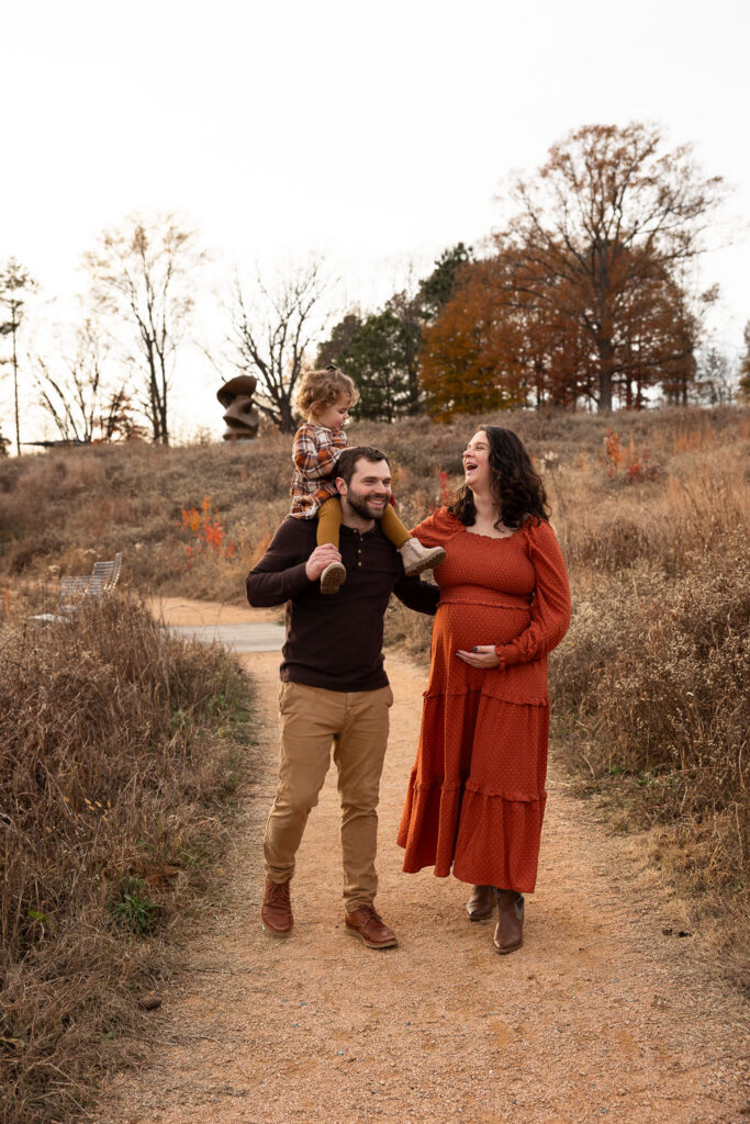 Raleigh maternity photo session at NCMA in the fall, family of three walks down natural path while mom cups her pregnant belly and dad carries toddler daughter on his shoulders