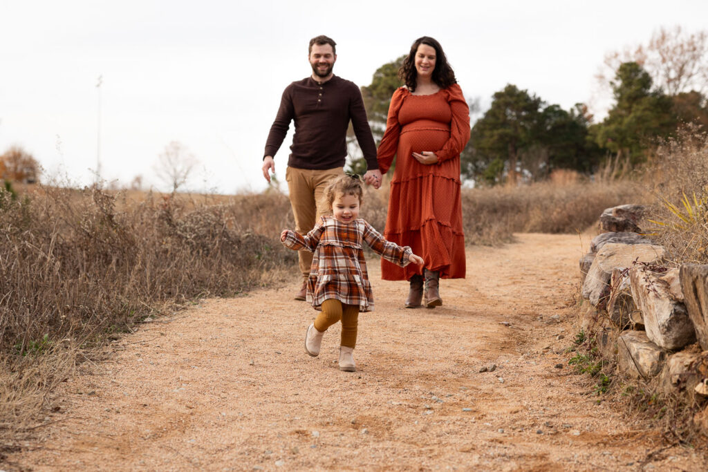Raleigh family of three maternity photos at NCMA, parents walking toward camera while toddler runs ahead